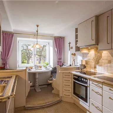 A modern kitchen with bright wood design and white tiles. A table with chairs stands in front of a window that lets in plenty of daylight.