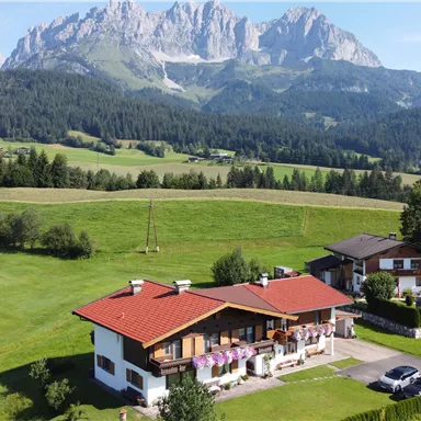 A picturesque landscape with green meadows and high mountains in the background. In the foreground, there are two cozy houses with red roofs.
