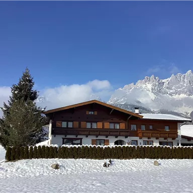A pretty wooden house in winter with a snow-covered ground. In the background, there are snow-capped mountains and a blue sky.