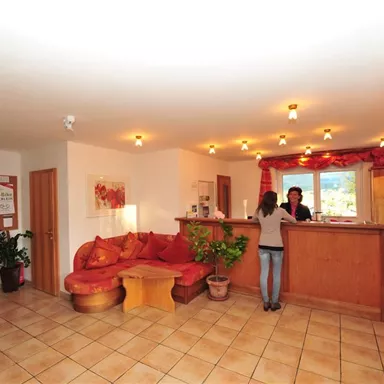 Ein Empfangsbereich mit einer großen Couch und einem Holzschalter. Es gibt Pflanzen und warmes Licht, was eine einladende Atmosphäre schafft.