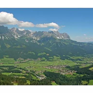 A stunning mountain landscape with green meadows and snow-capped peaks. The sky is clear and blue.