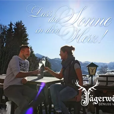 A couple sits at a table in the mountains and enjoys a drink in the sun. In the background, snowy mountains and pine trees can be seen.
