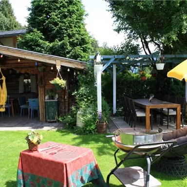An inviting garden with a romantic wooden cabin. There's a table, chairs, and a sunny sunshade.