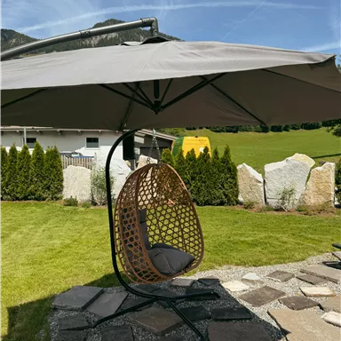 A cozy hanging chair under a sun umbrella in the garden. In the background, green meadows and large stones are visible.