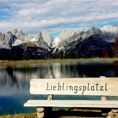 Eine Bank mit der Aufschrift "Lieblingsplatzl" steht am Ufer eines klaren Sees. Im Hintergrund erheben sich majestätische Berge unter einem blauen Himmel.
