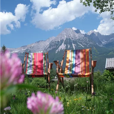 Two colorful deck chairs are standing in a green meadow with flowers. In the background, majestic mountains and a blue sky with some clouds are visible.