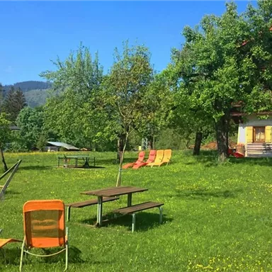 A green meadow with cozy loungers and shade-giving trees. In the background, there is a building with balconies and a view of the mountains.
