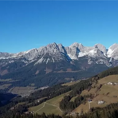 An impressive mountain landscape with snow-covered peaks and clear blue sky. Green meadows and small villages lie in the valleys.