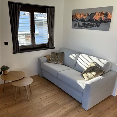 A cozy living room with a gray sofa and a small table. The sun is shining through the window onto the wooden floor.