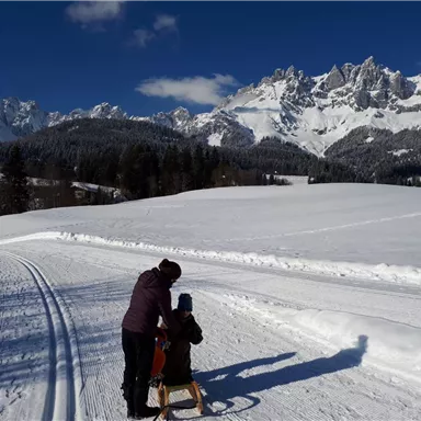 Eine winterliche Landschaft mit schneebedeckten Feldern und Bergen im Hintergrund. Ein Erwachsener hält die Hand eines Kindes, während sie den Weg entlang gehen.