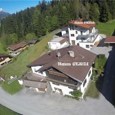 Ein ruhiges Wohngebiet mit mehreren Häusern, umgeben von Grünflächen und Bäumen. Im Hintergrund sind Berge und ein klarer Himmel zu sehen.