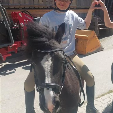 Ein Junge sitzt auf einem kleinen Pony und lächelt. Im Hintergrund sind Gebäude und Ausrüstungsgegenstände zu sehen.