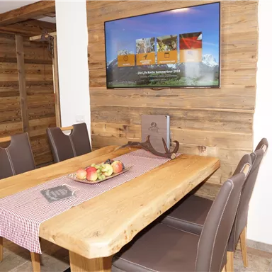 A cozy dining area with a large wooden table and modern chairs. A television is mounted on the wall, and a bowl of apples sits on the table.