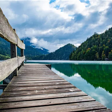 Ein Holzsteg führt über einen ruhigen See, umgeben von grünen Wäldern und Bergen. Der Himmel ist teilweise bewölkt und spiegelt sich im Wasser wider.