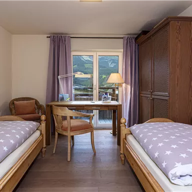 A cozy bedroom with two single beds and a desk. The room has a large window front that offers a view of the mountains.
