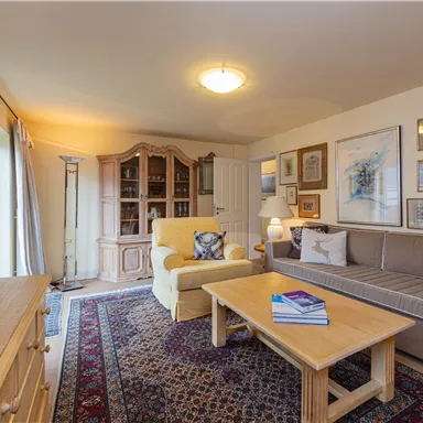 A cozy living room with a beige sofa and a yellow armchair. There is a wooden table, bookshelves, and decorative pictures on the walls.