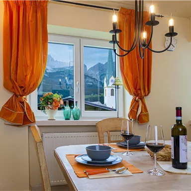 A cozy dining area with a wooden table, dishes, and wine. The view through the window shows the mountains and a church in the background.