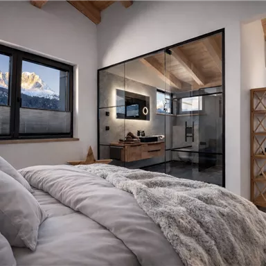 Ein modernes Schlafzimmer mit einem komfortablen Bett und einem Blick auf schneebedeckte Berge. Das Bad ist durch eine Glaswand getrennt und strahlt Eleganz aus.