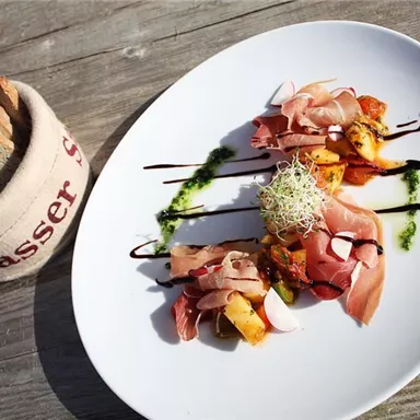 An artfully arranged plate with various types of ham and vegetables, garnished with sprouts. Next to the plate, there is a portion of bread in a basket.