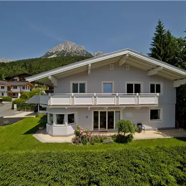 A modern house with a white facade and balcony, surrounded by a green meadow. In the background, mountains and forests are visible.
