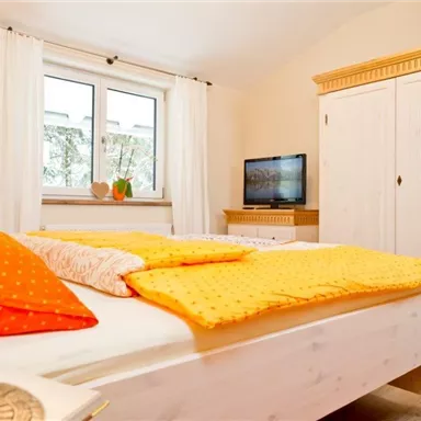 A bright bedroom with a large bed and colorful pillows. In the background, there is a television and a wardrobe.