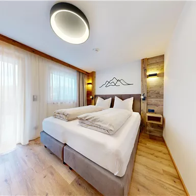 Ein modernes Schlafzimmer mit einem Doppelbett und hellen Vorhängen. Die warme Holzverkleidung und die sanfte Beleuchtung schaffen eine gemütliche Atmosphäre.