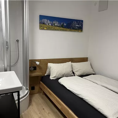 A small, modern room with a double bed and beige pillows. On the left side, there is a shower and a sink.