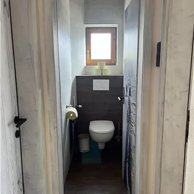 A small bathroom with a white toilet and a window. The walls are painted in neutral tones and the floor is dark.