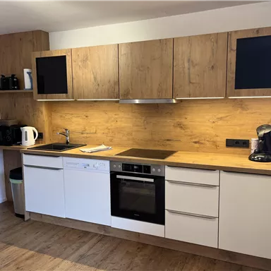 A modern kitchen with wooden shelves and white cabinets. The equipment includes a kettle, a coffee machine, and a dishwasher.