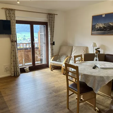 A cozy living space with a dining table, armchairs, and a television on the wall. A beautiful view of the mountains is visible through the window.