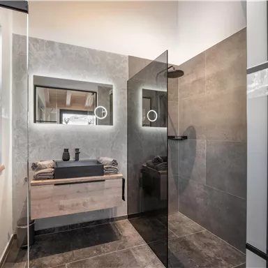 Ein moderner Badezimmer mit eleganten Fliesen und einer Glasdusche. Eine minimalistische Holzkommode und stilvolle Beleuchtung runden das Design ab.