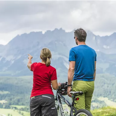 Ein Paar steht auf einem Hügel und betrachtet die Berge. Sie haben Fahrräder dabei und genießen die Aussicht.
