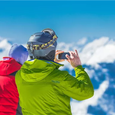 A skier in a green jacket is taking a photo of snow-covered mountains. In the background, a clear blue sky is visible.