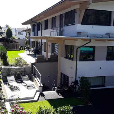A modern building with a balcony and spacious terrace. The garden is well-kept and furnished with loungers and plants.