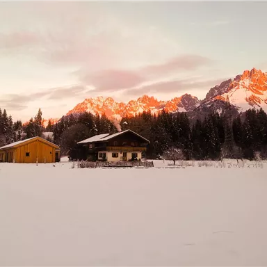 A picturesque winter landscape with snow-covered fields and wooden houses. In the background, the mountains shine in the sunlight.