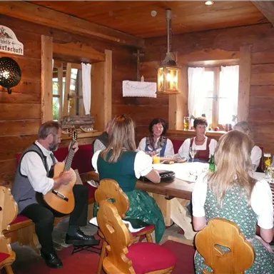 A cozy room with wooden decor and traditional Bavarian costumes. People are sitting at the table enjoying music and camaraderie.