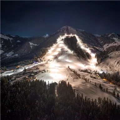 A wintry mountain landscape at night with illuminated ski slopes. In the foreground, snow-covered trees are visible.
