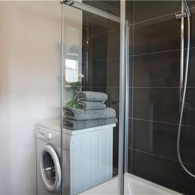 A modern bathroom with a shower and a washing machine. Piles of gray towels are stacked on the washing machine.