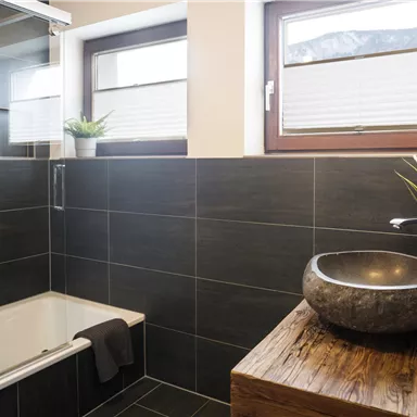A modern bathroom with dark tiles and an elegant stone sink. The large shower and the windows provide plenty of light.