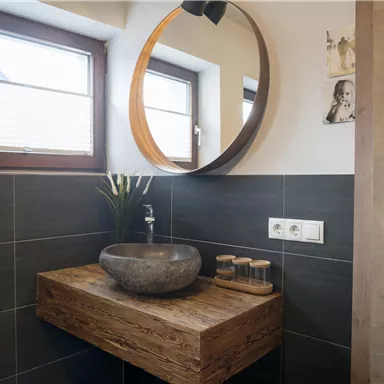 A modern bathroom with a large round mirror and a stylish stone sink. The walls are tiled in dark colors, and there are some decorative elements like plants.