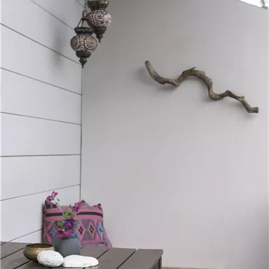 A stylish outdoor area with a wooden table and colorful cushions. A decorative lamp and a natural wooden branch hang on the wall.