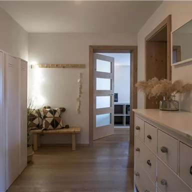 A bright hallway with wooden flooring and modern furnishings. On the left side, there are cabinets and a bench, while on the right side, there is a dresser.