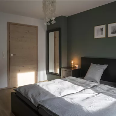 A modern bedroom with a large bed, a mirror, and stylish artworks on the wall. The room has a warm atmosphere due to the sun rays that fall in.
