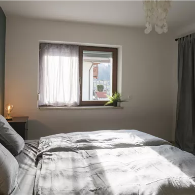 A bright bedroom with a double bed and soft pillows. Next to the window are plants, and the walls are painted in neutral colors.