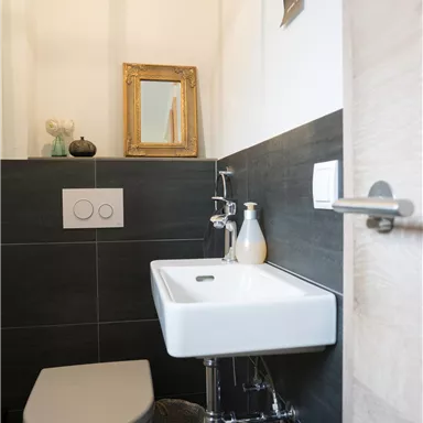 A modern bathroom with a stylish toilet and a small sink. The walls are in a dark tone and there are decorative elements such as a mirror and a bottle.