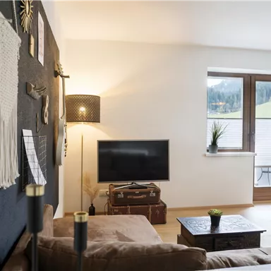 A cozy living room with a dark wall, a TV, and a window overlooking greenery. The decor is modern and inviting, with a sofa and a table.