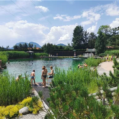 Ein Naturbad mit klaren Wasser und viel Grün. Menschen genießen die Sonne und die Umgebung.