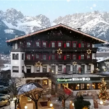 A traditional hotel with festive Christmas decorations in a wintry landscape. In the background, majestic mountains and a clear sky can be seen.