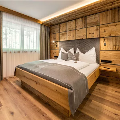 A cozy bedroom with a wooden bed and modern furniture. The walls are clad in wood, and large windows provide plenty of daylight.