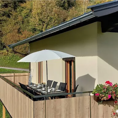 Ein schöner Balkon mit einem Esstisch und Stühlen, geschützt durch einen Sonnenschirm. Umgeben von grünen Bäumen und Blumen.